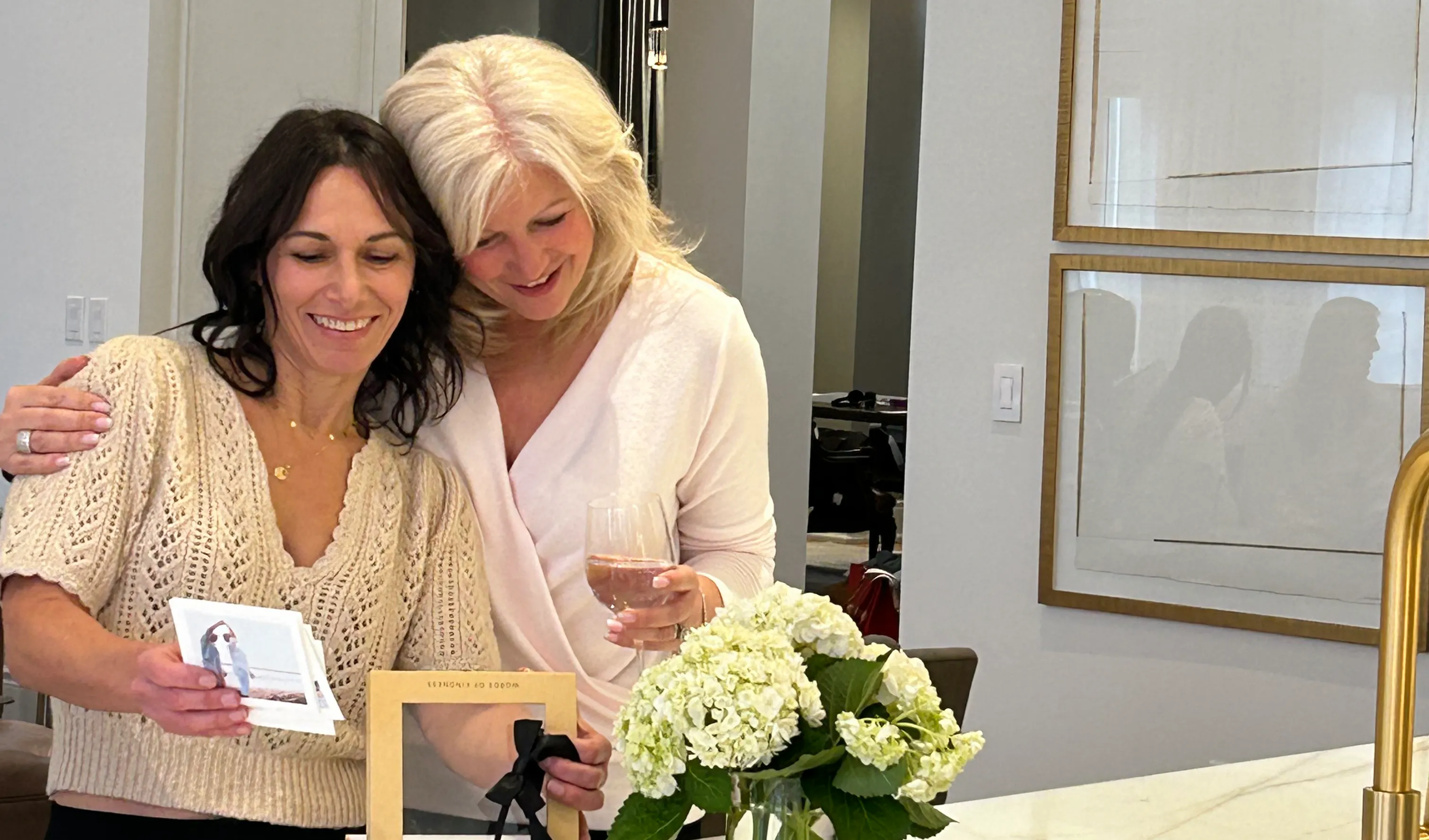 Two women sharing a warm hug in a bright kitchen while reading heartfelt message cards and photos from an open Woxbox keepsake box tied with a black ribbon