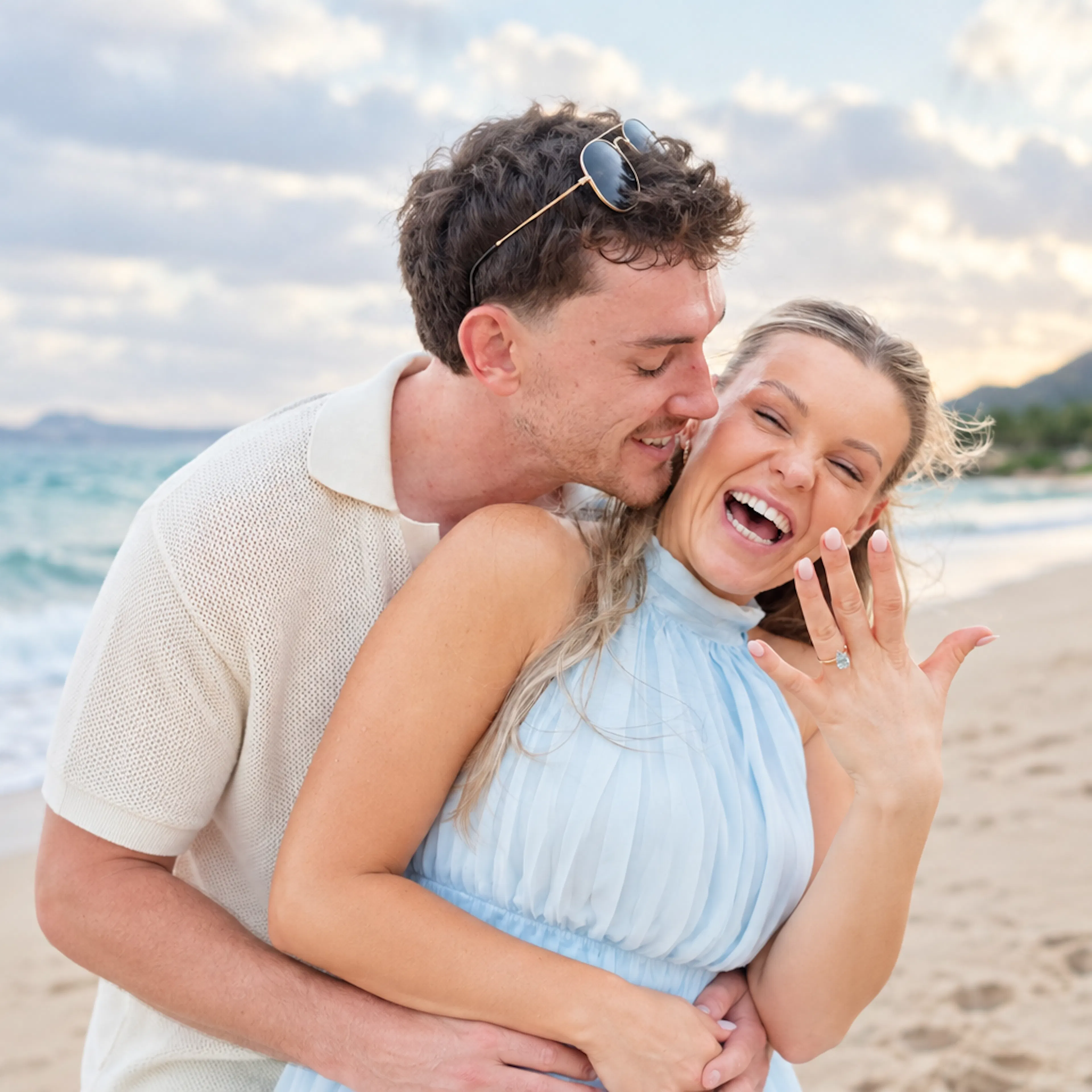 Newly engaged couple laughing and embracing as she shows off her engagement ring on a beach