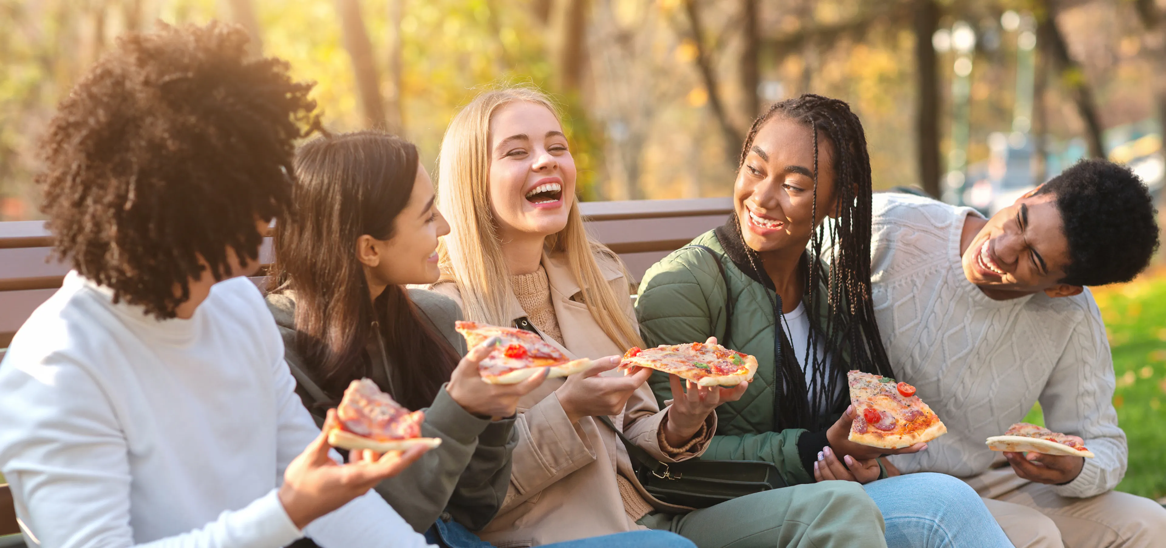 Group of friends laughing and sharing pizza outdoors on a fall day, reconnecting in real life without their phones