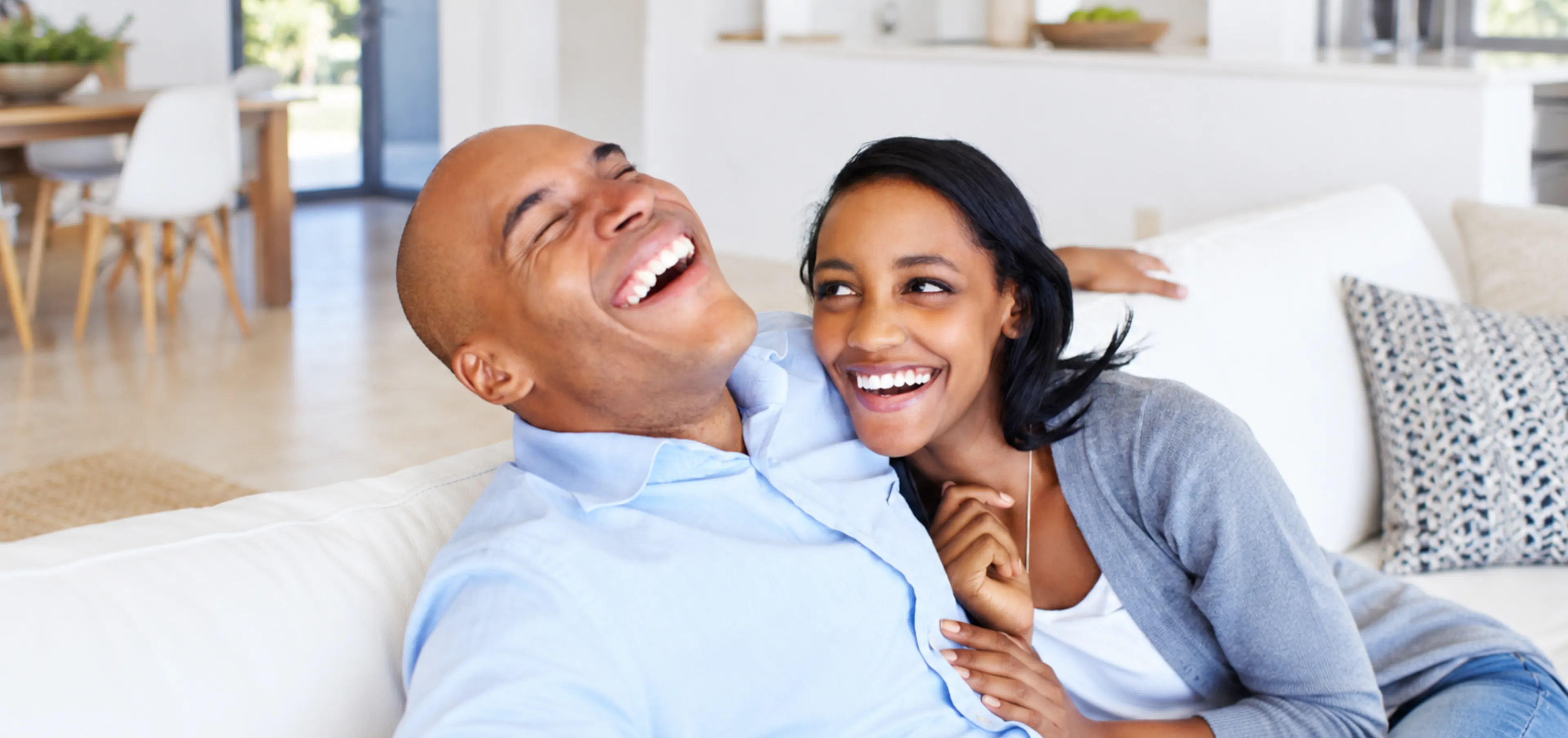 Happy couple laughing together on a white couch at home, sharing a lighthearted moment of connection