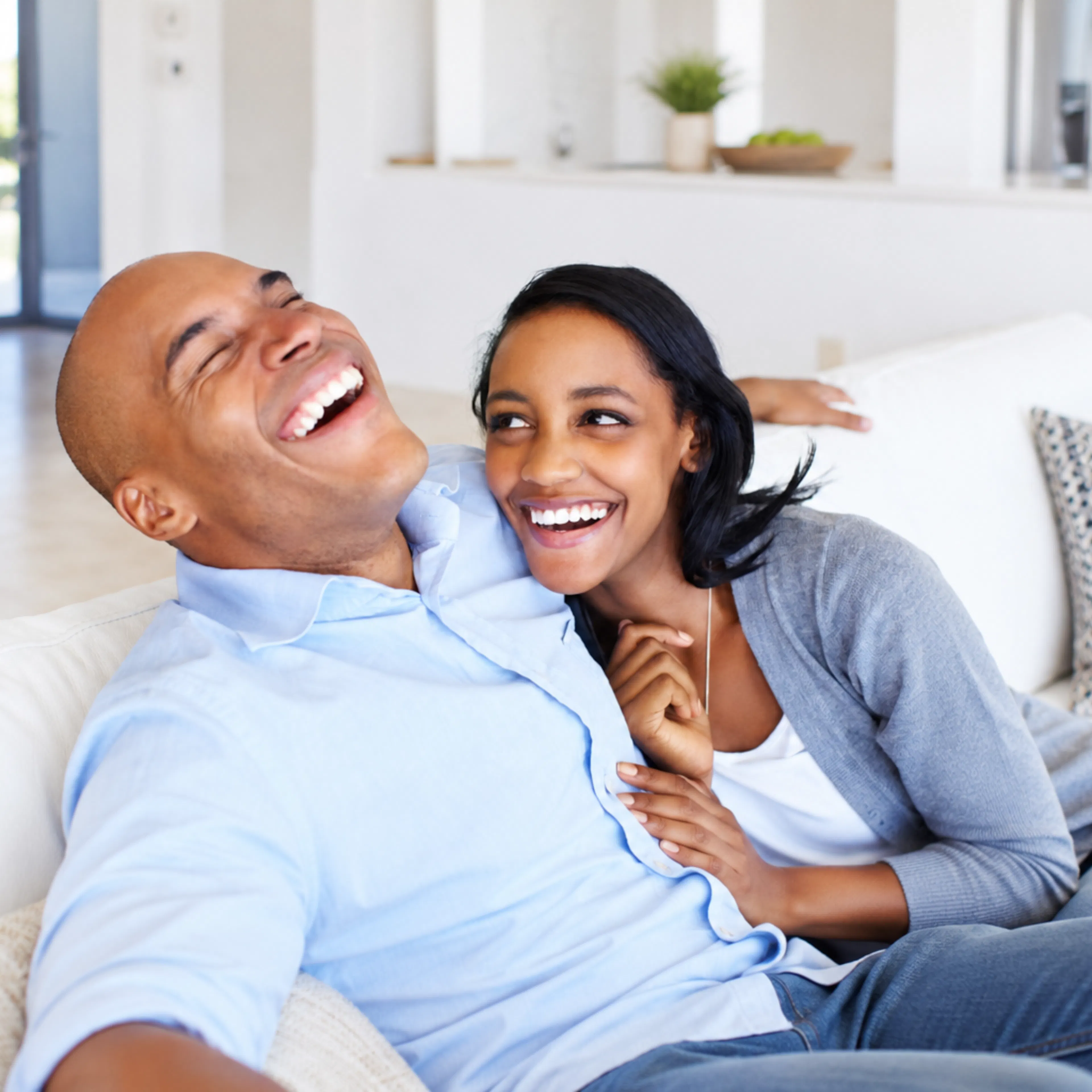 Happy couple laughing together on a white couch at home, sharing a lighthearted moment of connection