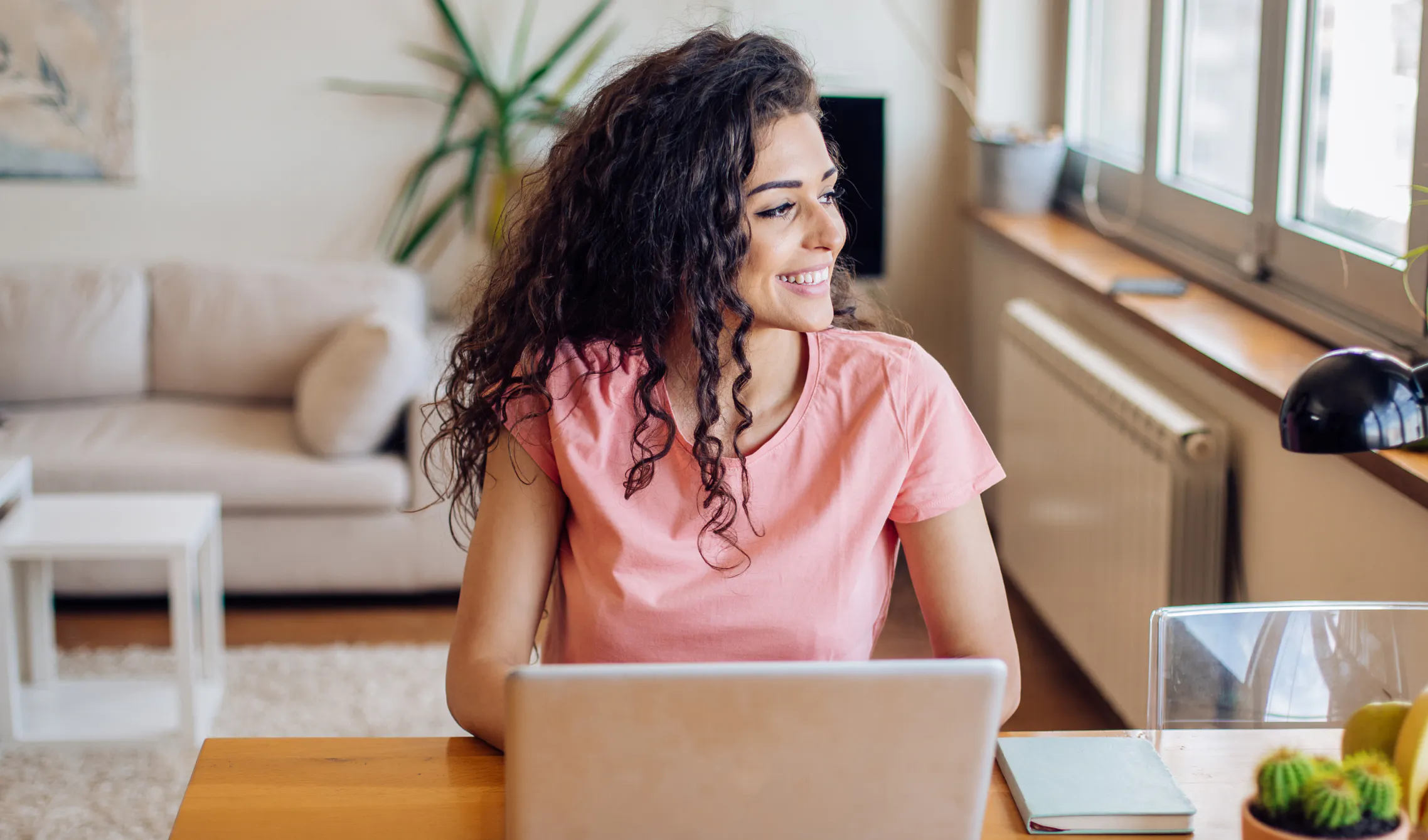 Woman smiling and gazing out a sunny window beside her laptop and notebook, embracing a get-to mindset at work
