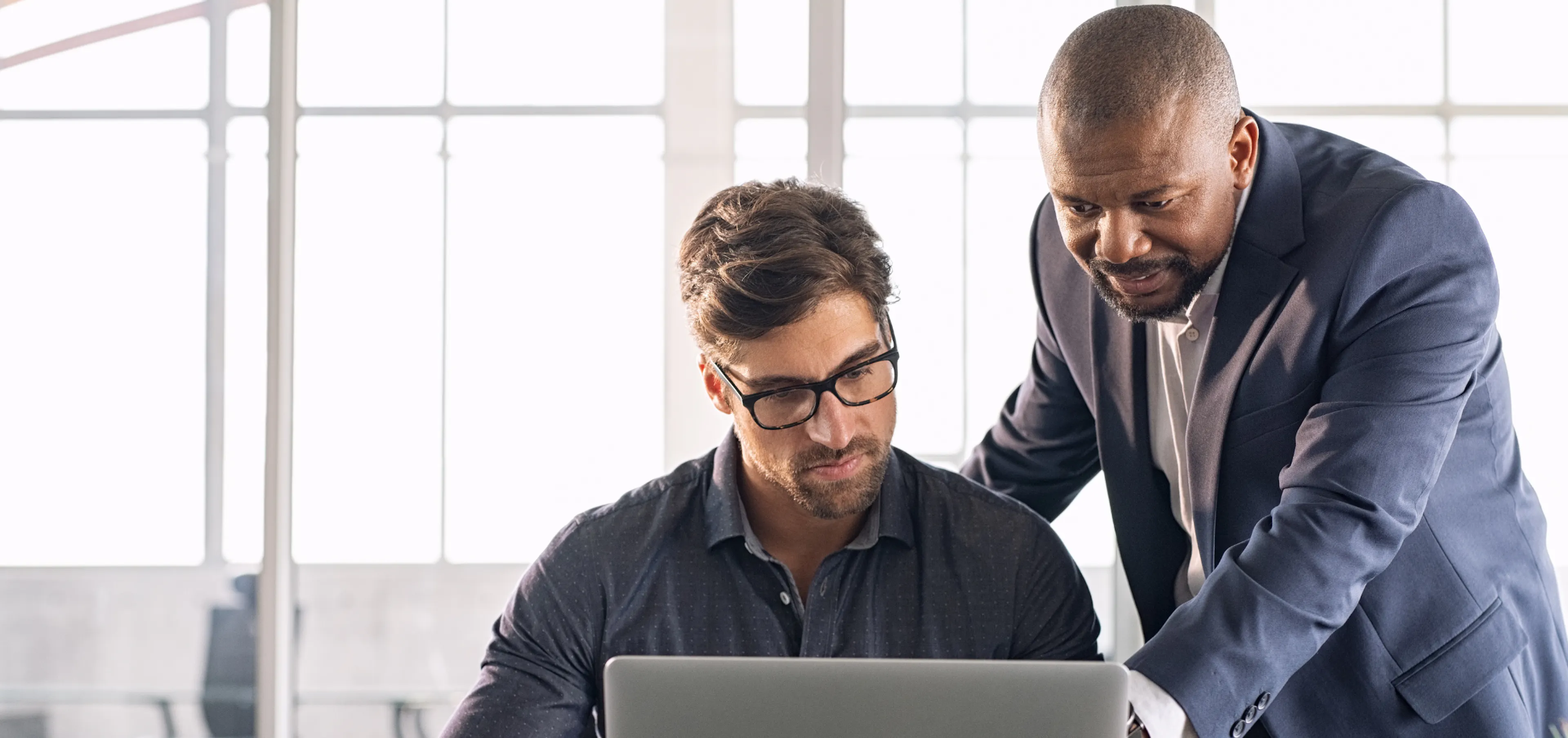 Manager leaning over an employee's laptop to review his work, noticing the effort behind the scenes