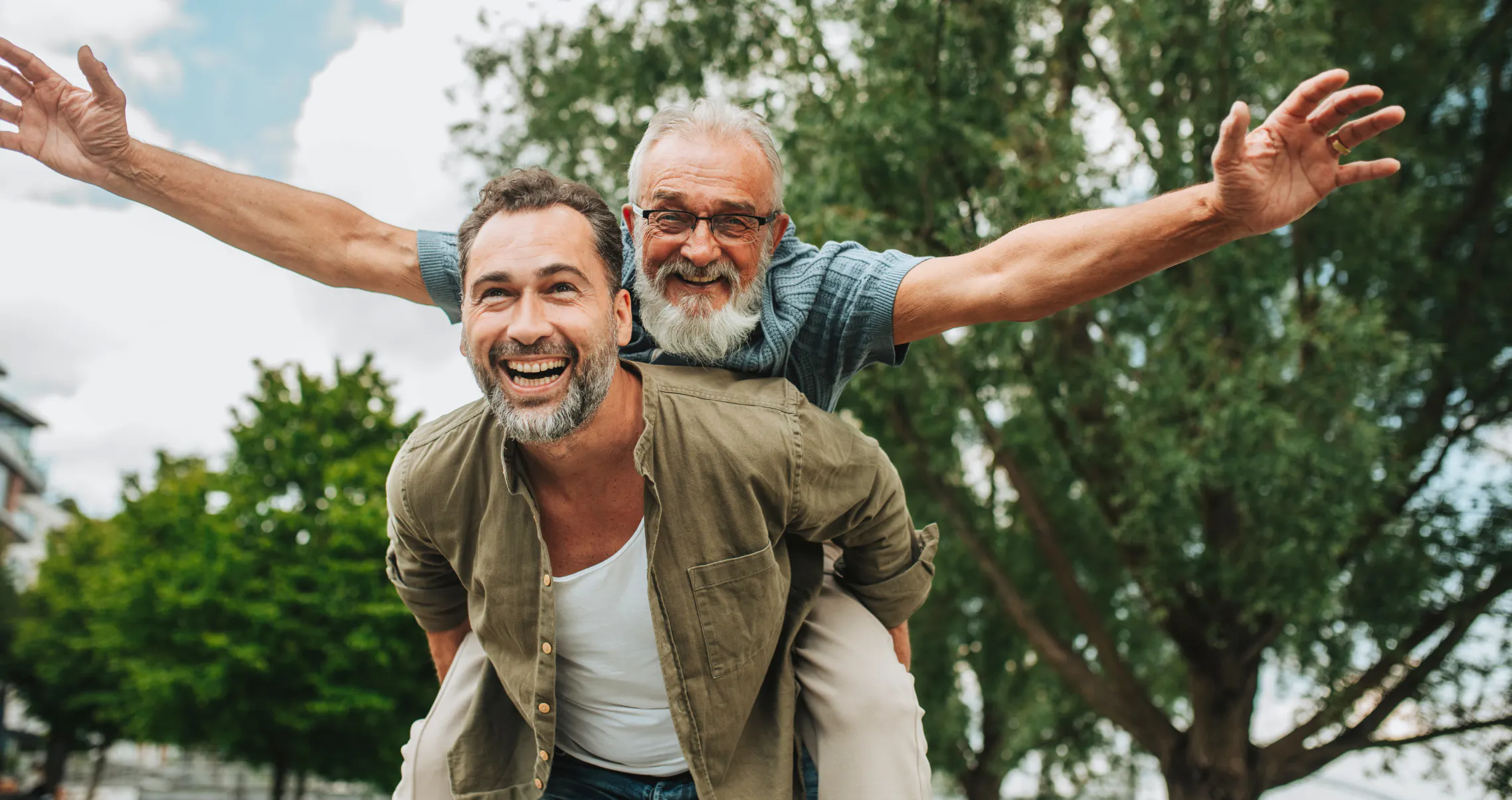 Middle-aged man laughing as he gives an older man with a white beard a piggyback ride in the park, celebrating intergenerational friendship