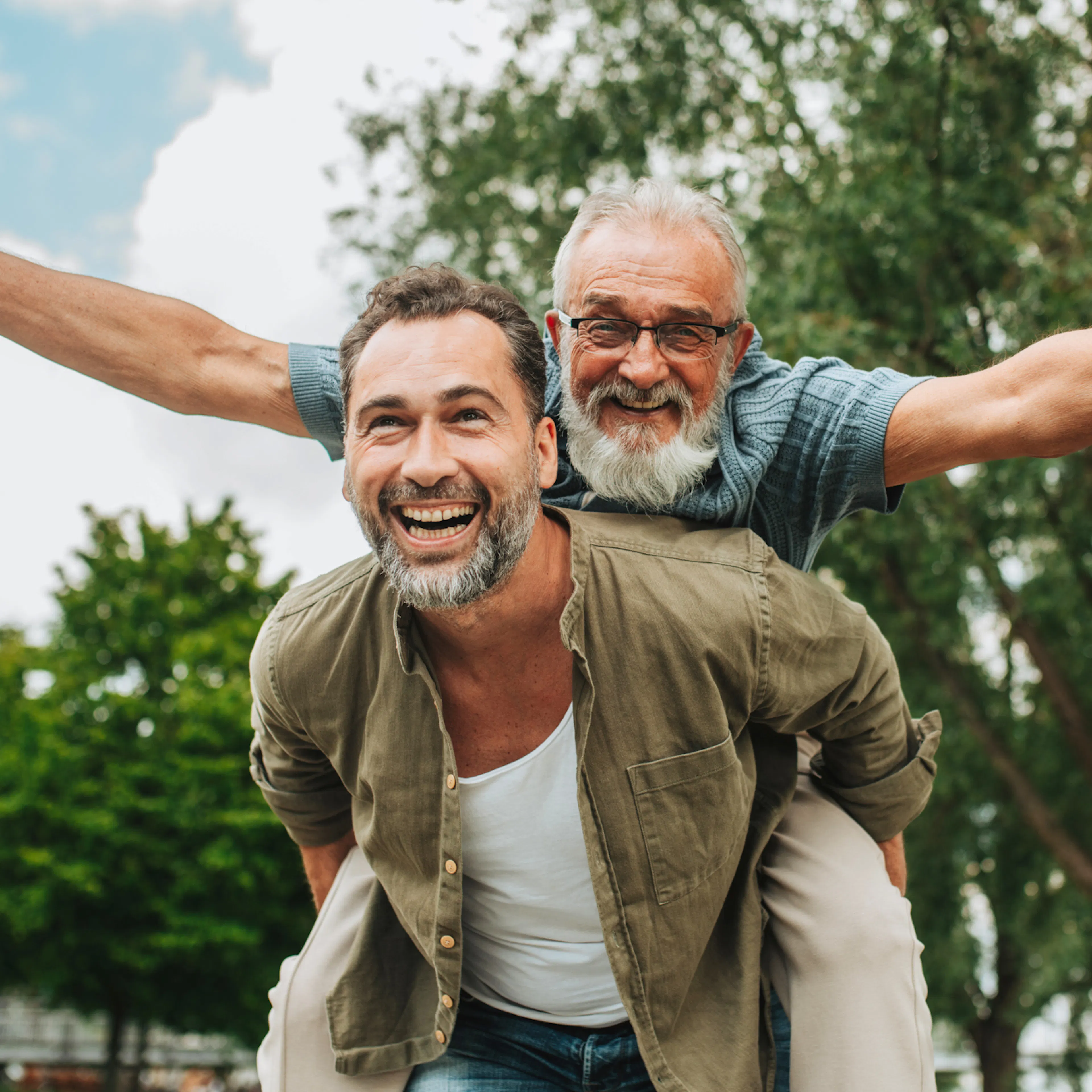 Middle-aged man laughing as he gives an older man with a white beard a piggyback ride in the park, celebrating intergenerational friendship