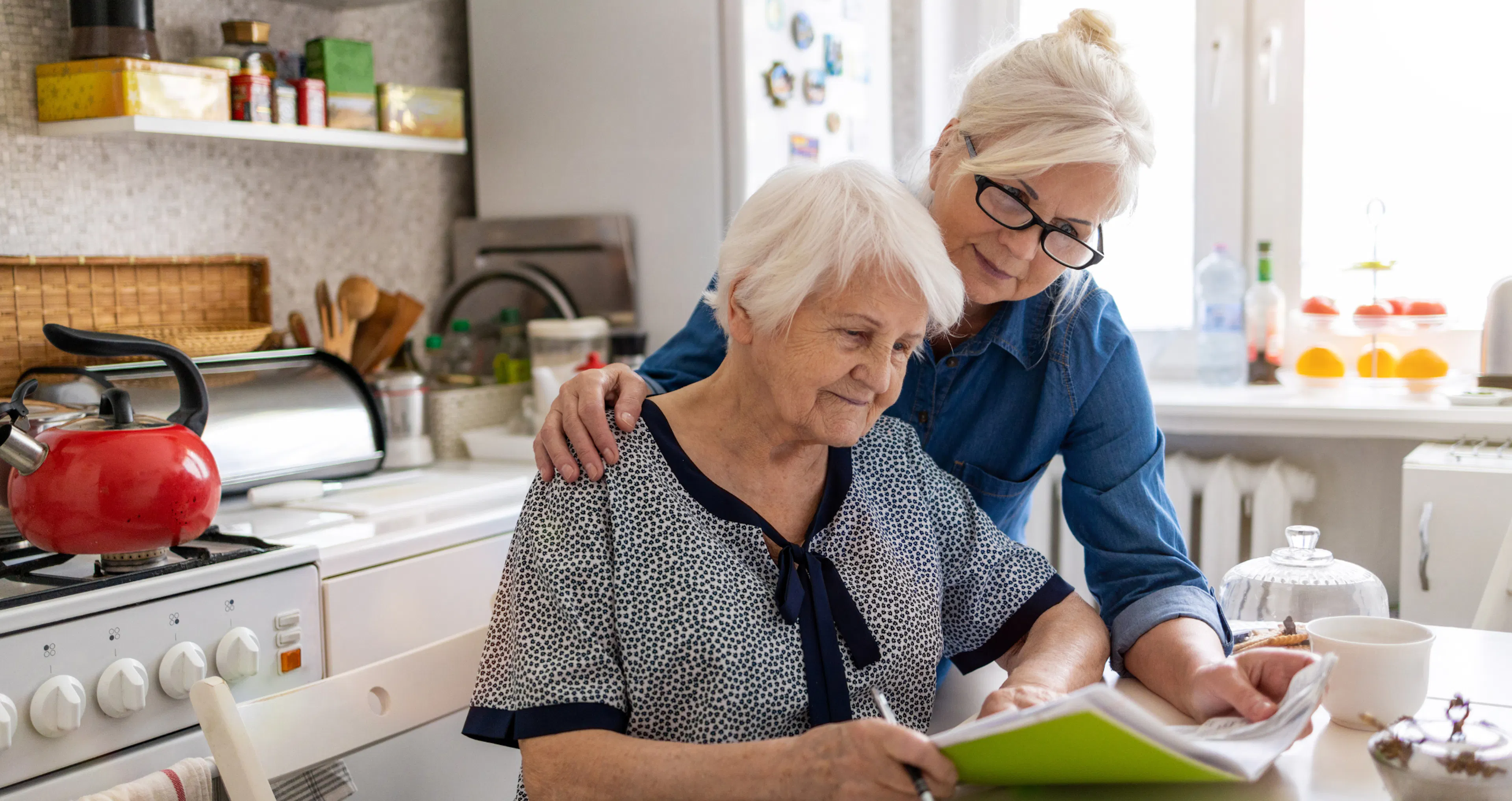 Daughter leaning in with a hand on her elderly mother’s shoulder as they go over a notebook together at the kitchen table