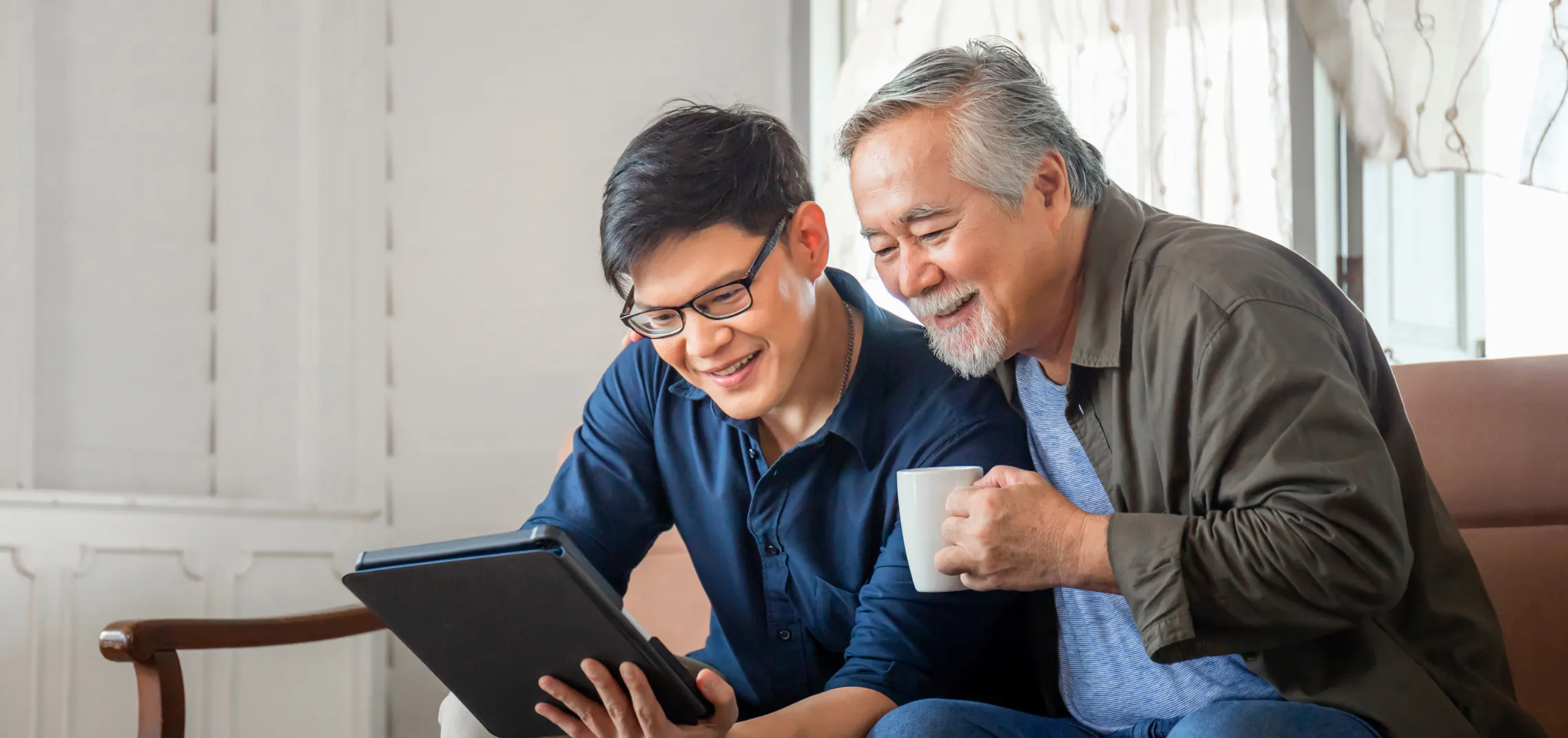 Younger man showing an older man something on a tablet as they share coffee and a laugh on the couch