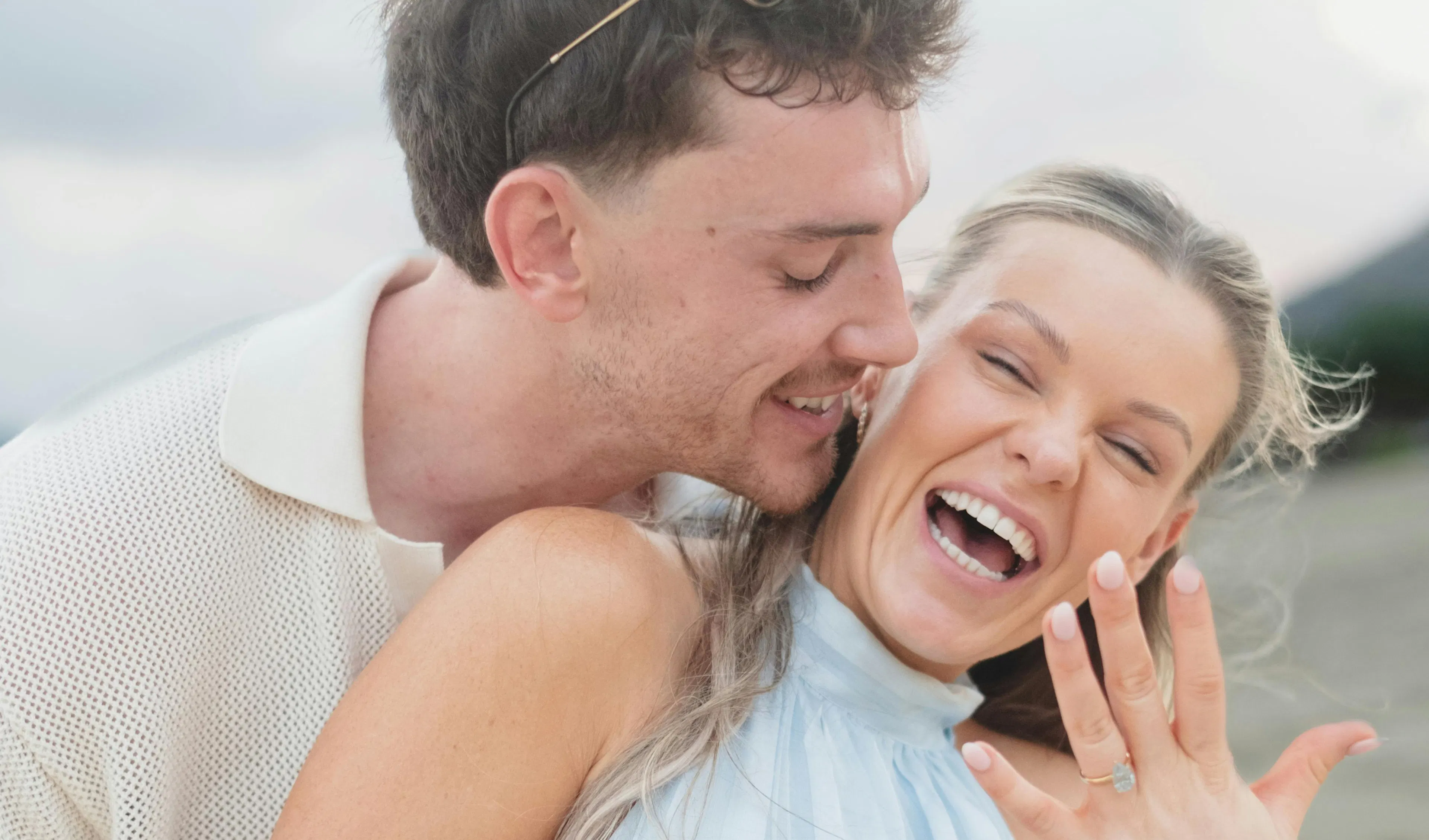 Newly engaged couple laughing and embracing as she shows off her engagement ring on a beach