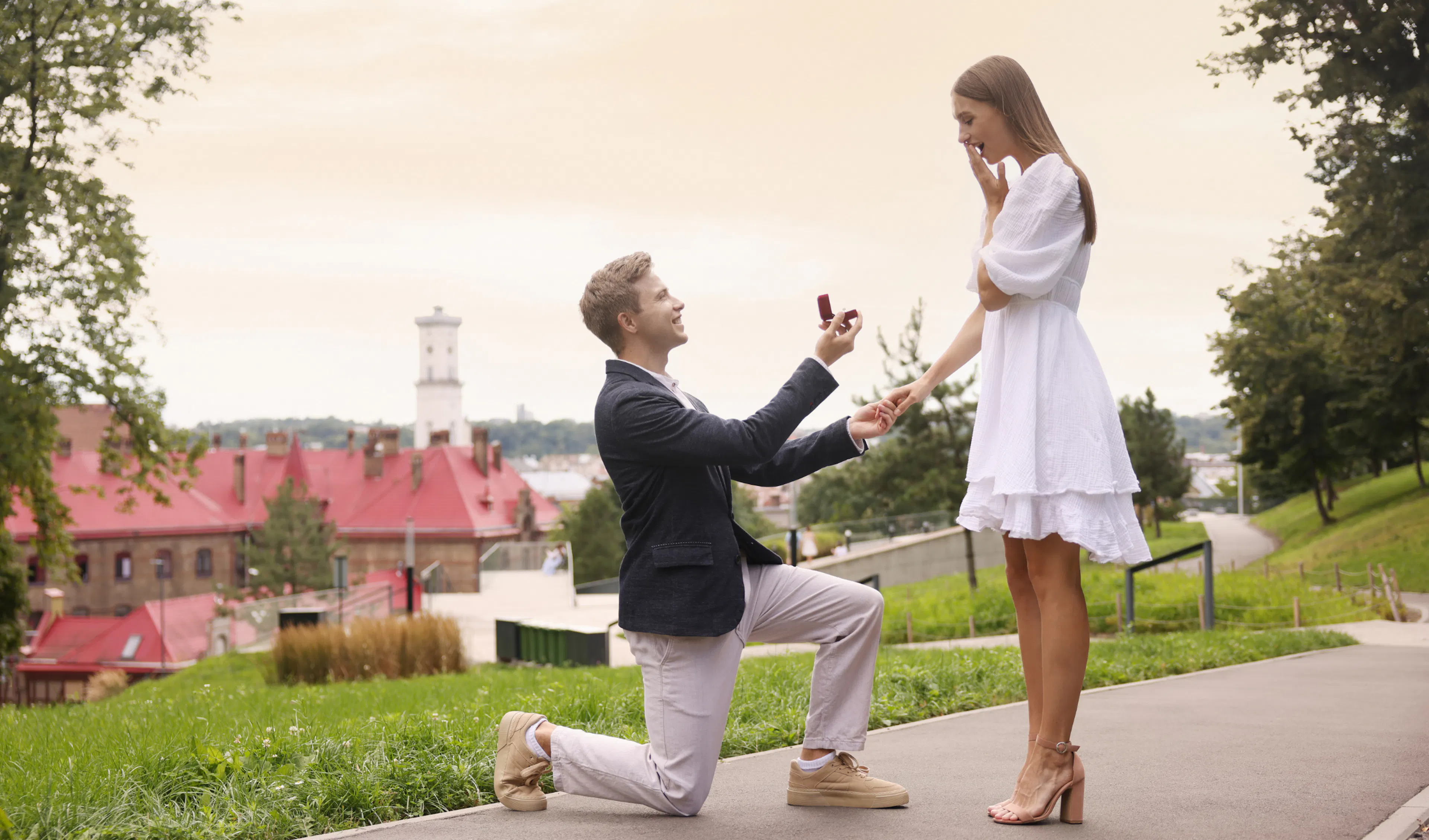Man kneeling with a ring box as a surprised woman in a white dress covers her mouth during a park proposal