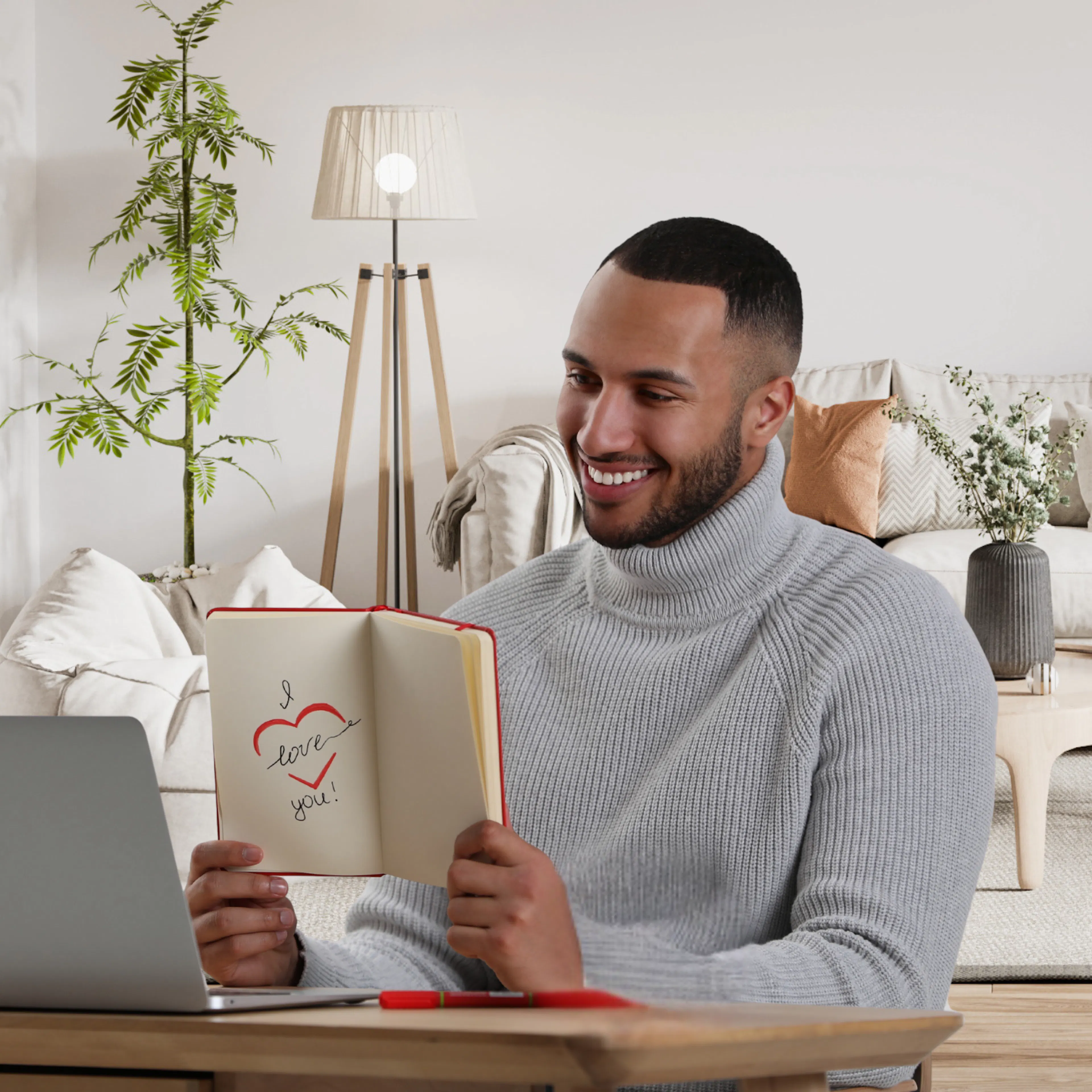 A man at his desk holding up a book that says "I love you", smiling at a video call on his laptop, representing a long-distance relationship.