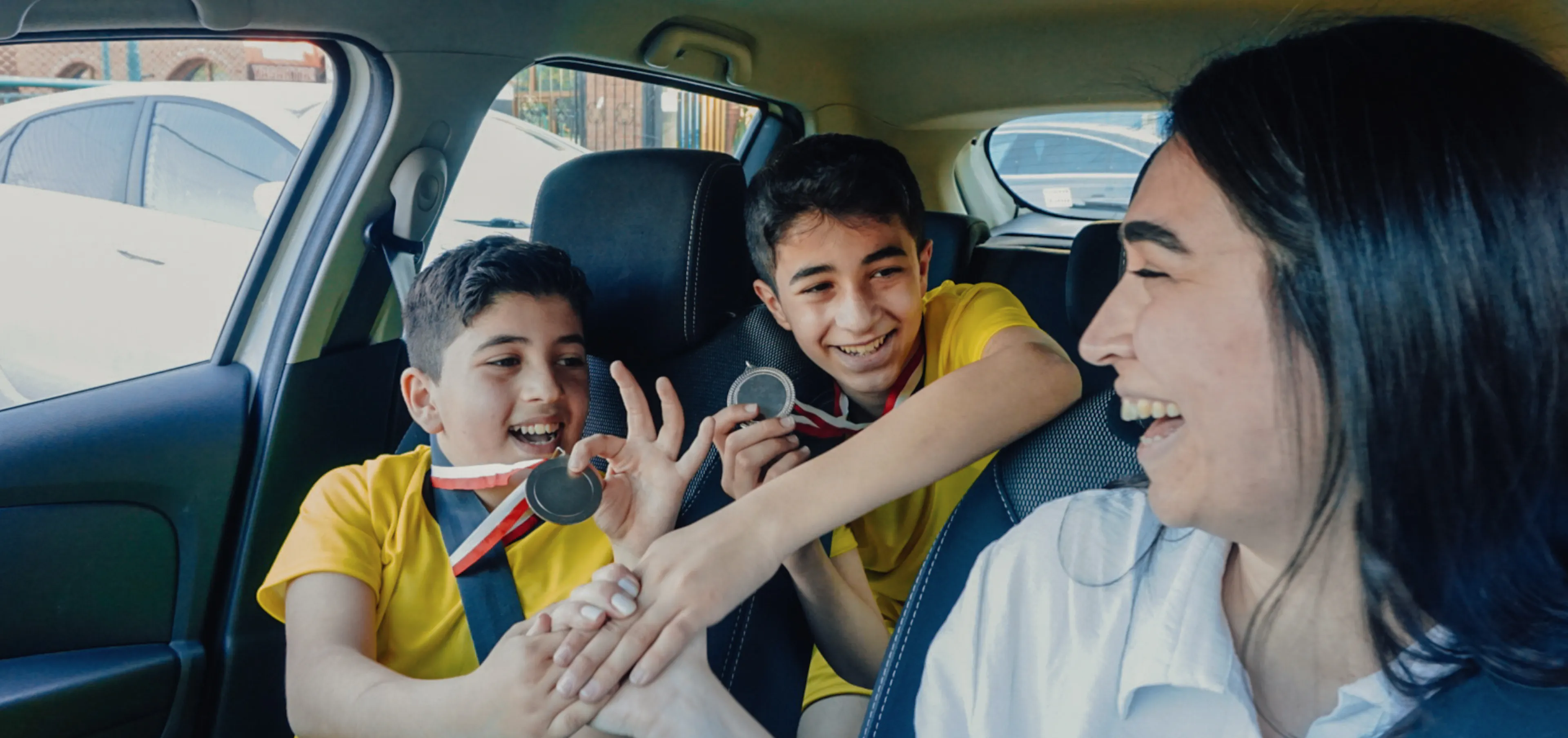 Mom laughing with two boys wearing sports jerseys and medals in the car, enjoying the ride home from practice