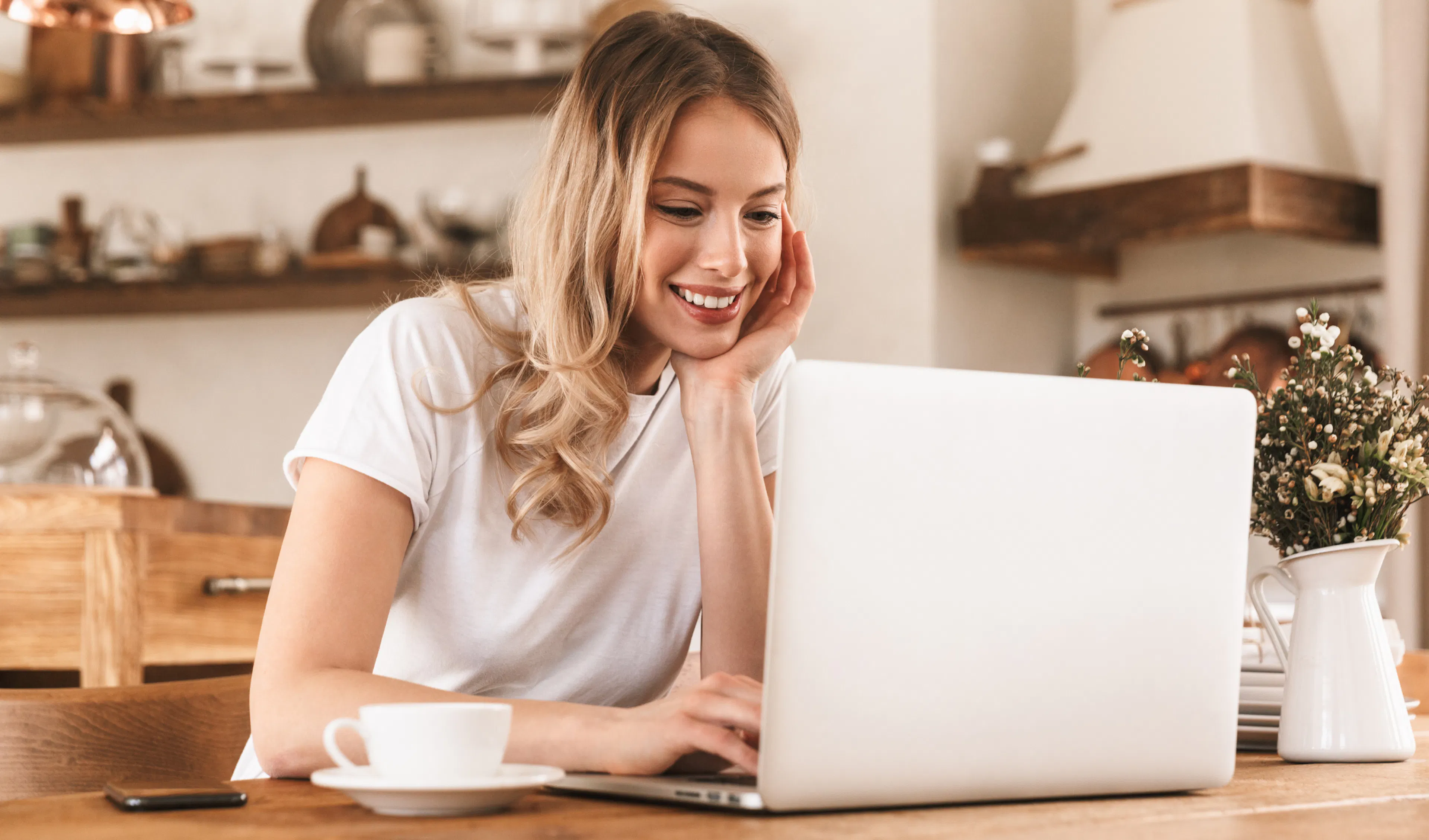 Woman smiling at a video call on her laptop in the kitchen, with a warm mug in evening light