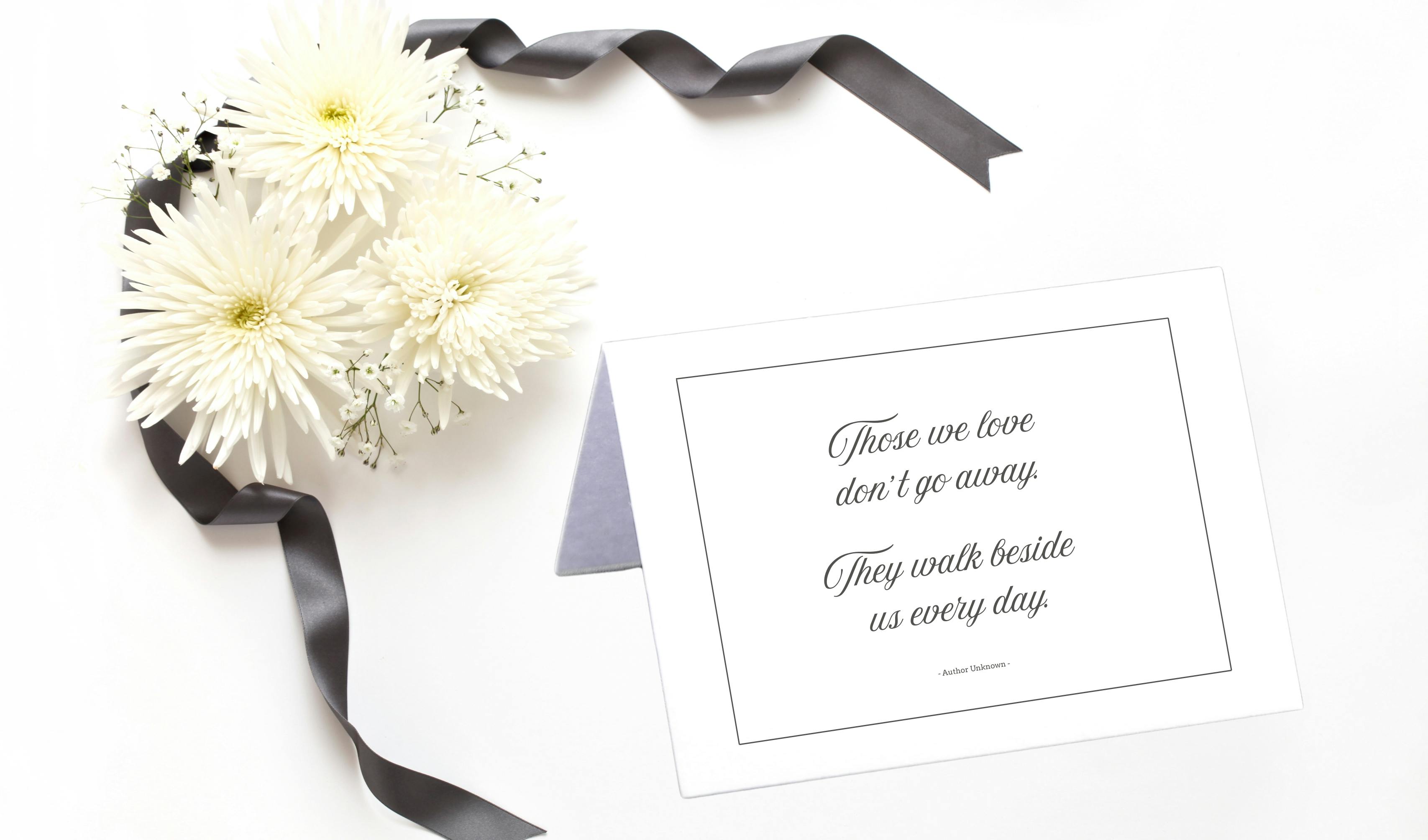 Close-up of fresh flowers and condolence card with a quote that reads "Those we love don't go away. They walk beside us every day. Author Unknown"