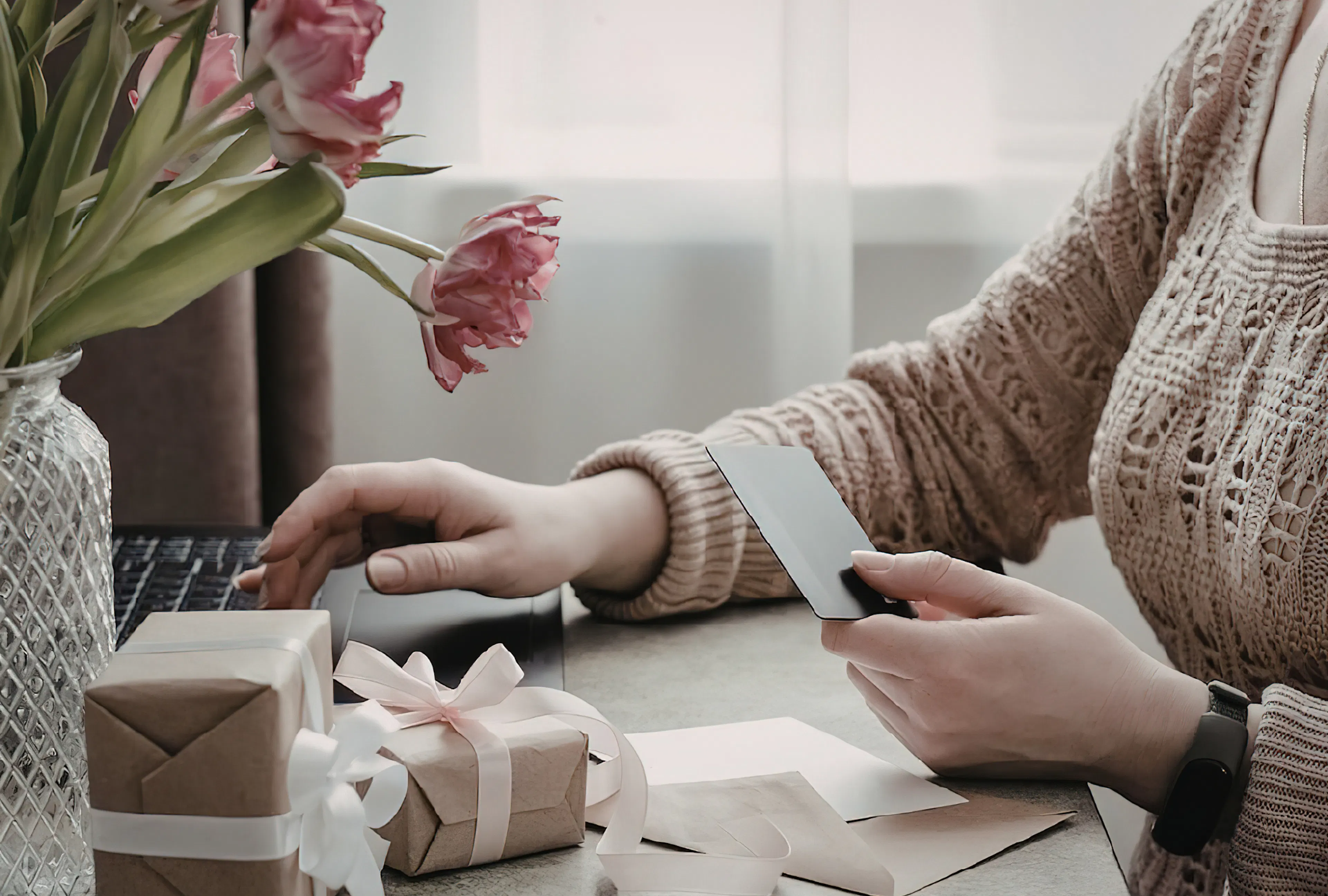A person writing a heartfelt message in a greeting card at a desk with wrapped gifts and flowers