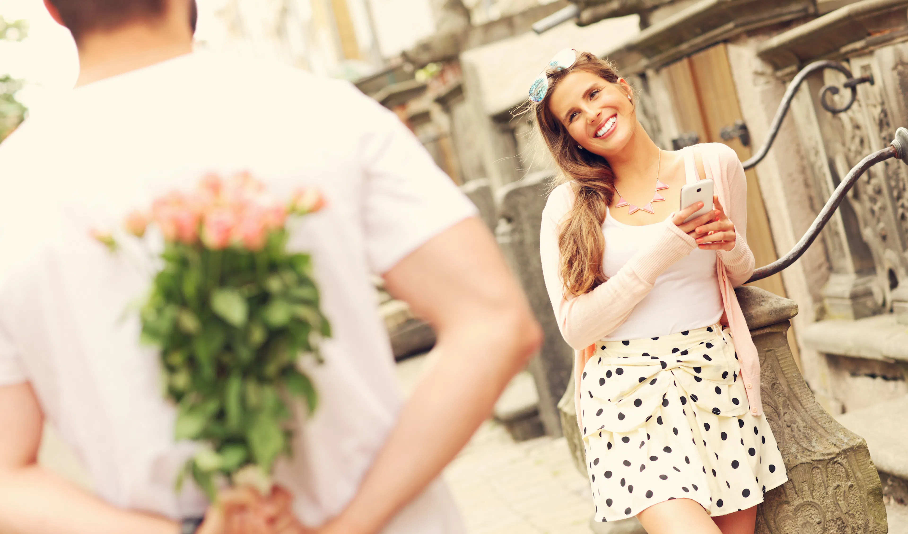 Smiling woman looking at her phone as her partner hides a bouquet of pink roses behind his back