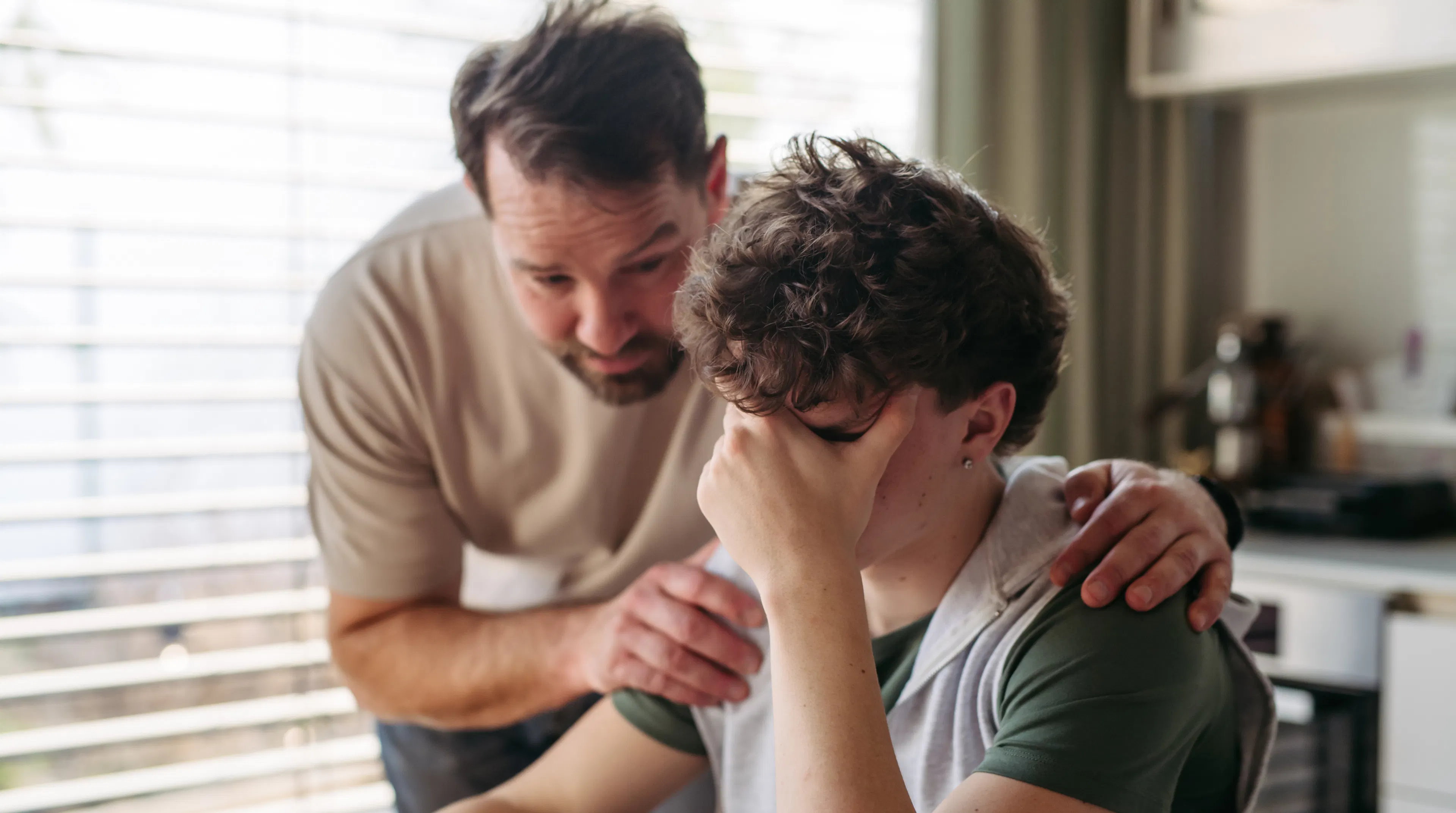 Father comforting son who is struggling with mental health, sitting in the kitchen with his hand covering his face
