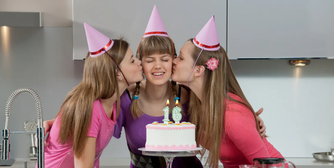 Young girl standing in front of her cake and smiling while her two best friends give her a kiss on the cheek for her sweet 16 birthday