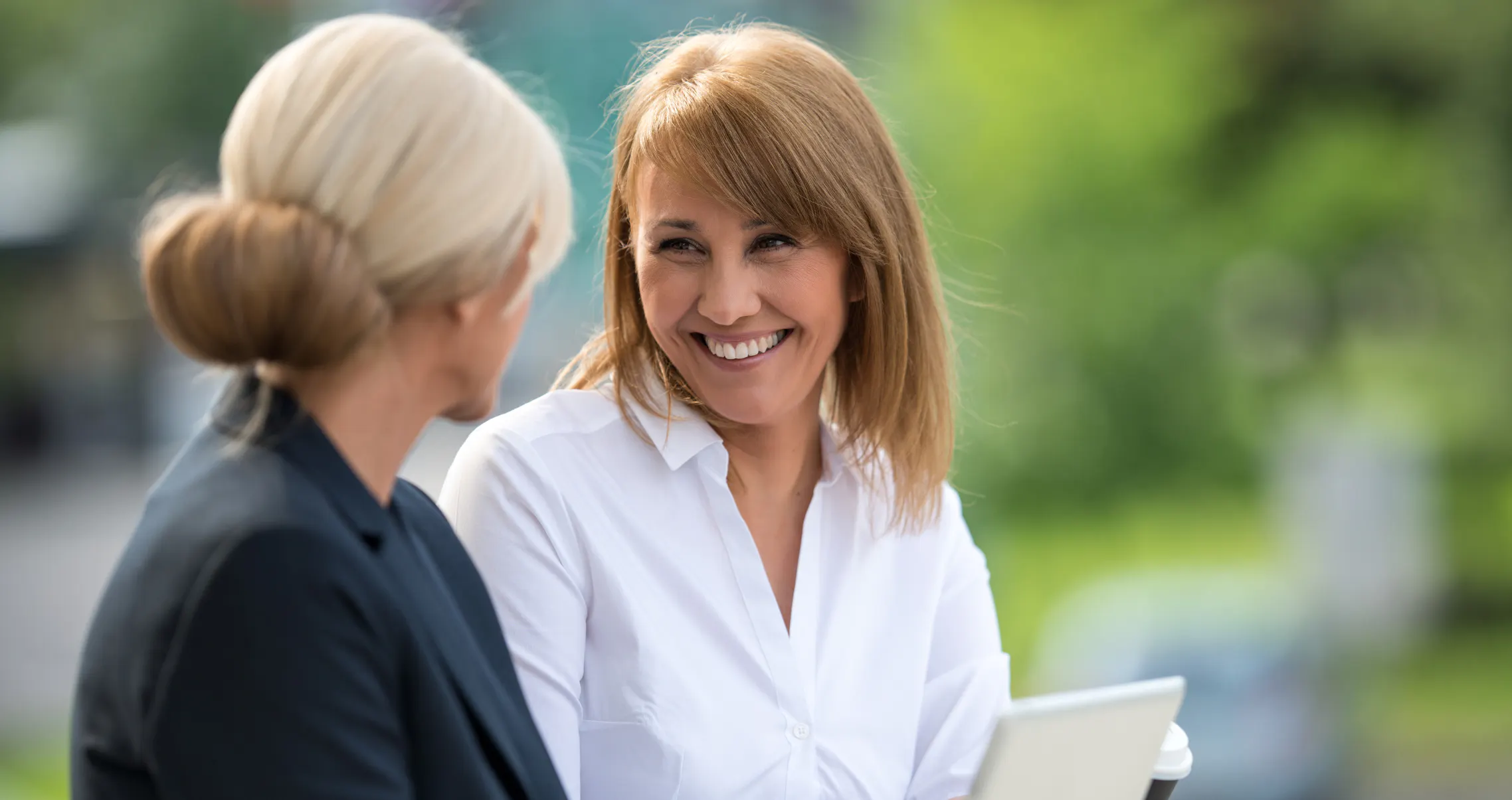 Smiling woman sitting outdoors with a colleague, looking pleased as her coworker shares a few words of appreciation