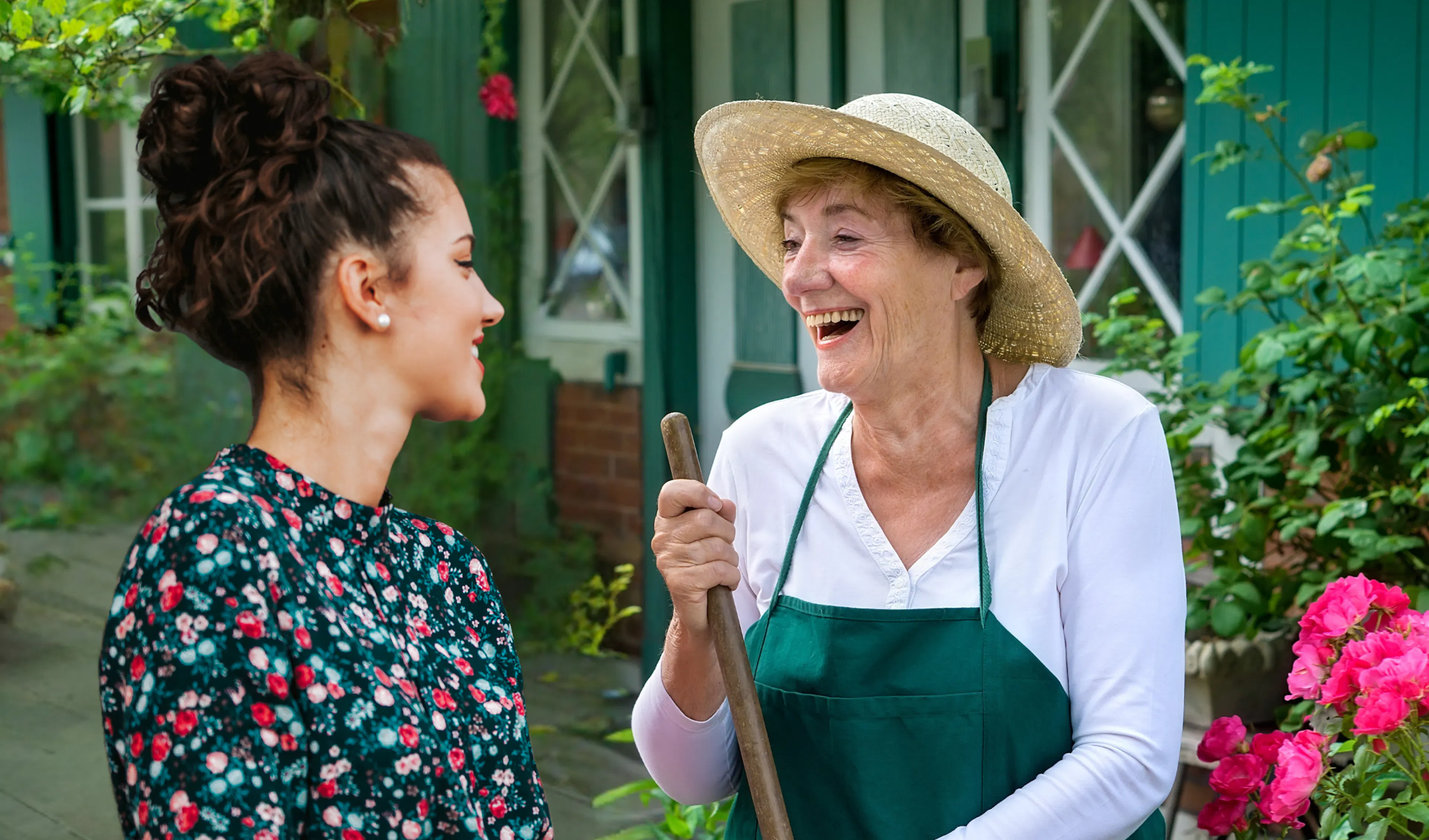 Older woman in a straw hat and gardening apron laughing with a younger neighbour outside her home, an example of making friends of different ages