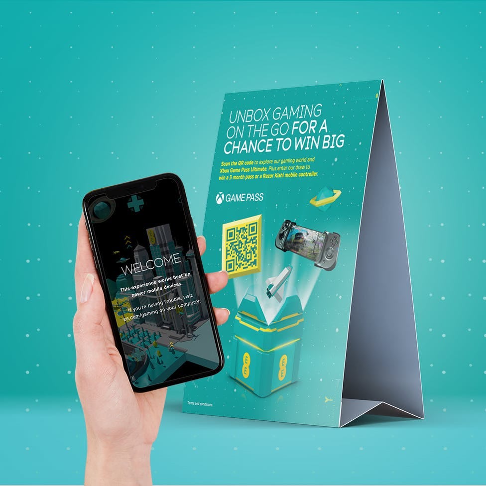 Hand holding a phone with a welcome screen to an application. In the background a display card advertising EE's Unbox Gaming on the Go experience.