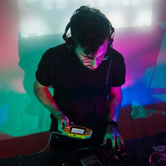 2xAA on stage - a man wearing headphones holding a Game Boy Advance while operating a mixing desk