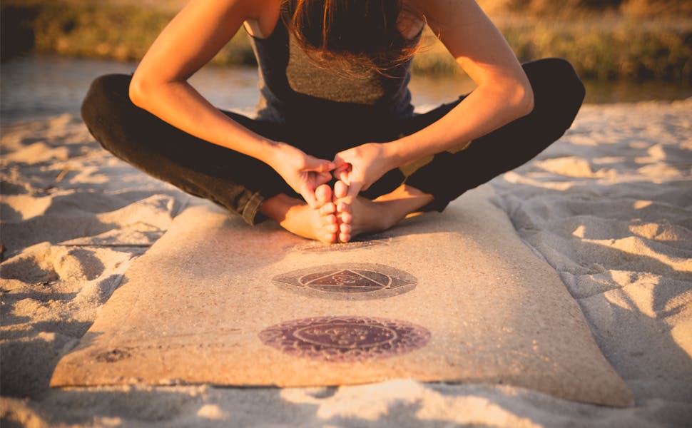 A woman does yoga on a cork mat on a sand beach.