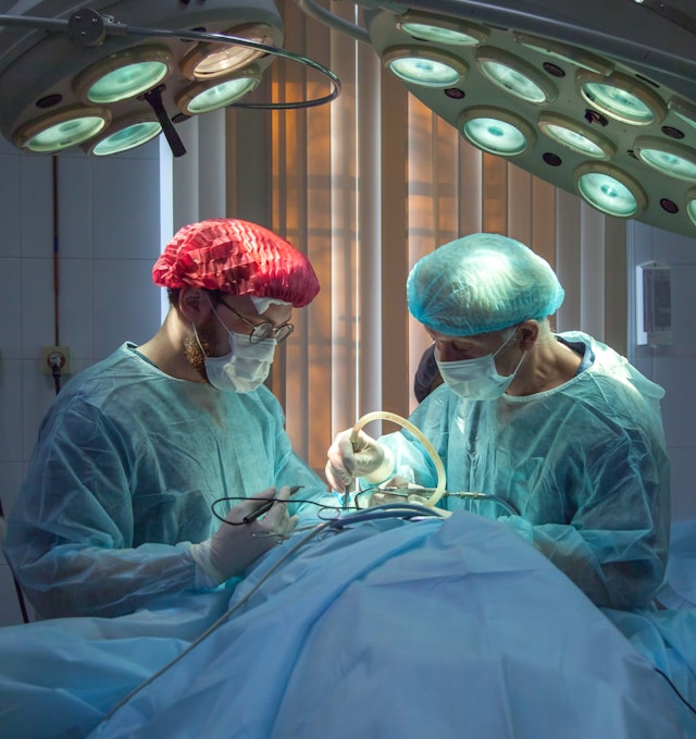 Two surgeons in blue scrubs, wearing masks and hairnets, are performing a surgical procedure on a patient under bright operating room lights.