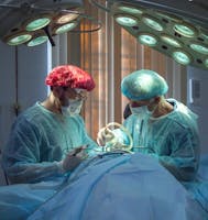 Two surgeons in blue scrubs, wearing masks and hairnets, are performing a surgical procedure on a patient under bright operating room lights.