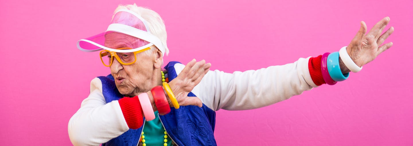 Elderly lady dressed in bright colours and jewellery and a pink hat is dabbing.