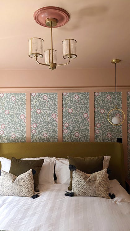 Close-up of a hanging ceiling light above a bed that sits in front of pink panelled wall with Briar Rose - Salix wallpaper.