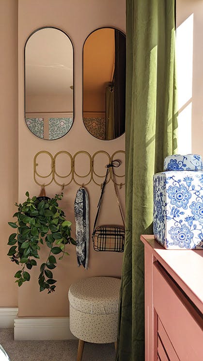 Close-up of a bedroom corner with pink walls, two oval mirrors, hooks, a small stool and a green curtain framing the window.