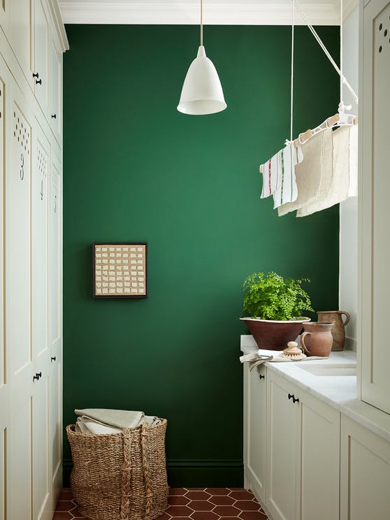 Laundry room painted in dark green shade 'Dark Brunswick' with a laundry basket, white cupboards and tiled floor.