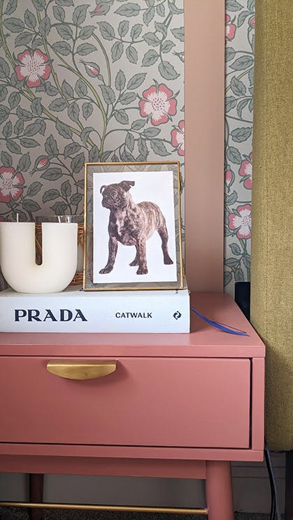 Close-up of a pink bedside table decorated with a book, candle and frames image in front of Briar Rose - Salix wallpaper.