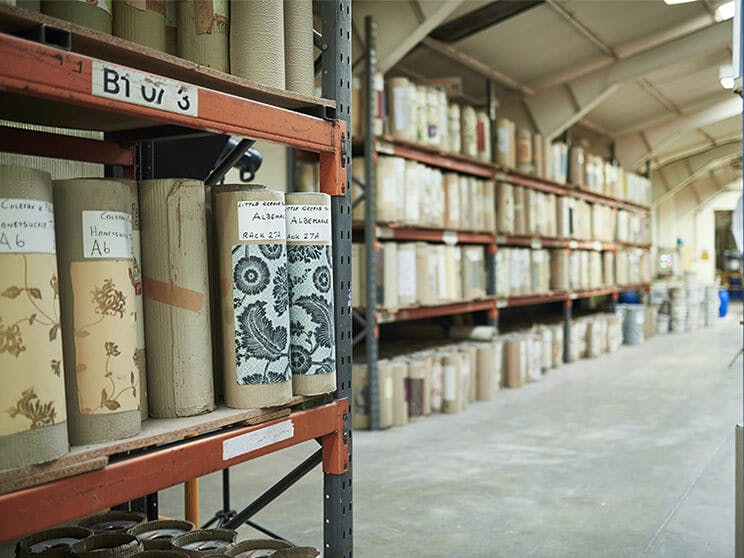 Warehouse shelves storing rolls of labelled Little Greene wallpapers wrapped in protective paper.