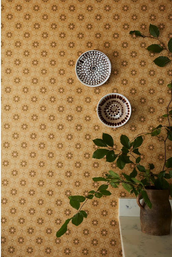 Close up of the golden brown small print floral wallpaper 'Ditsy Block - Bombolone' on a wall with a leafy plant on the right and wall plates.