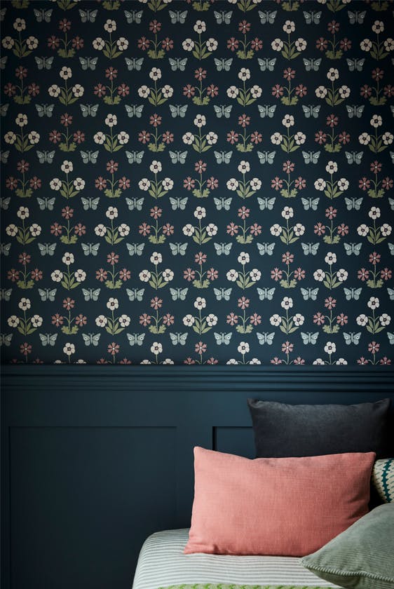 Close up of the dark blue small print wallpaper 'Burges Butterfly - Hicks' Blue' on a bedroom wall with co-ordinating paleing on the lower wall and a bed with cushions.