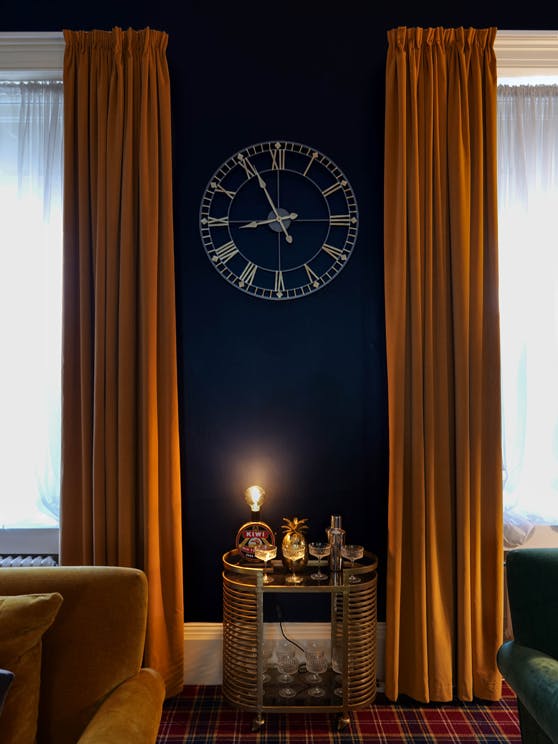 Sitting area with dark Dock Blue wall featuring a white clock in between two windows framed with warm yellow curtains.