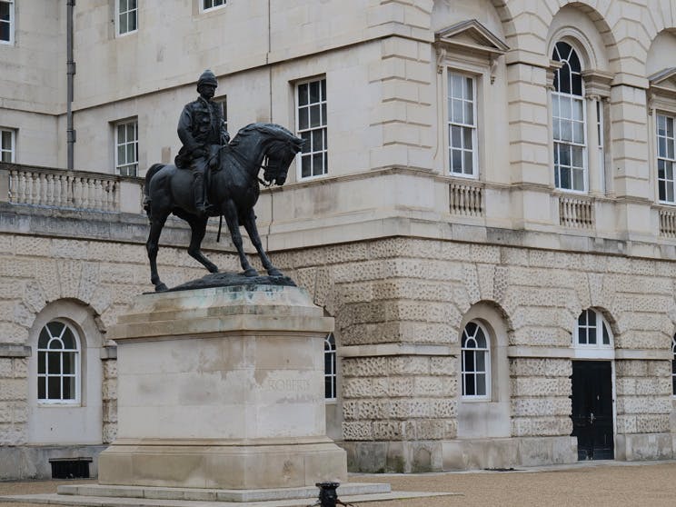 Black statue of a man riding a horse in front of the beige stone eighteenth-century Horse Guards building in London.