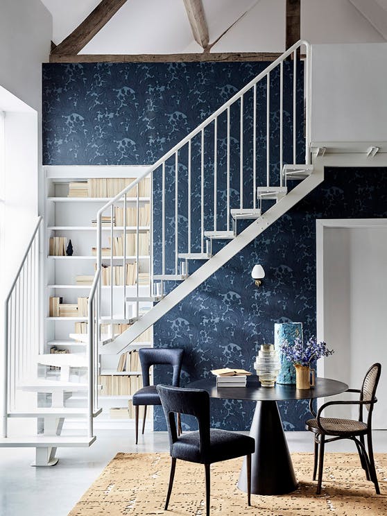 Hallway with dark blue damask wallpaper (Tulip - Blue Black), a black table and chairs, and white stairs passing a bookcase.