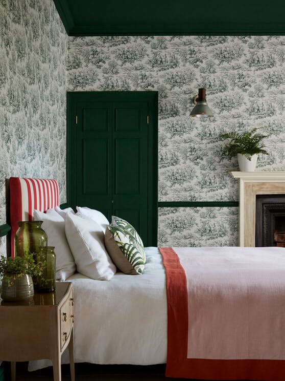 Bedroom with dark green French Toile wallpaper (Lovers' Toile - Puck), dark green (Puck) with a lamp and plant on a desk.