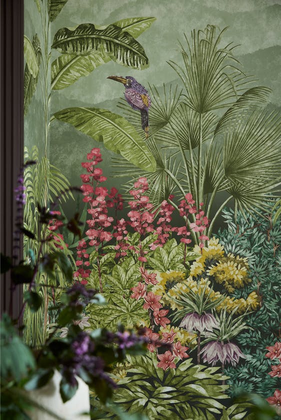 Close up of the vibrant green tropical mural wallpaper 'Capricorn - Boringdon' on a wall with a leafy plant in the foreground. 