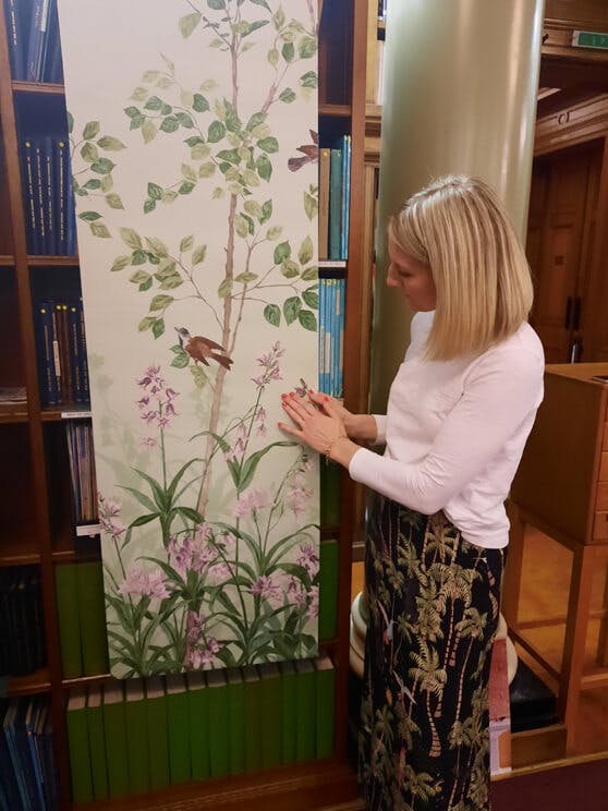 Woman (Ruth) looking at a strip of the Bird & Bluebell wallpaper design placed in front of a wooden bookcase.