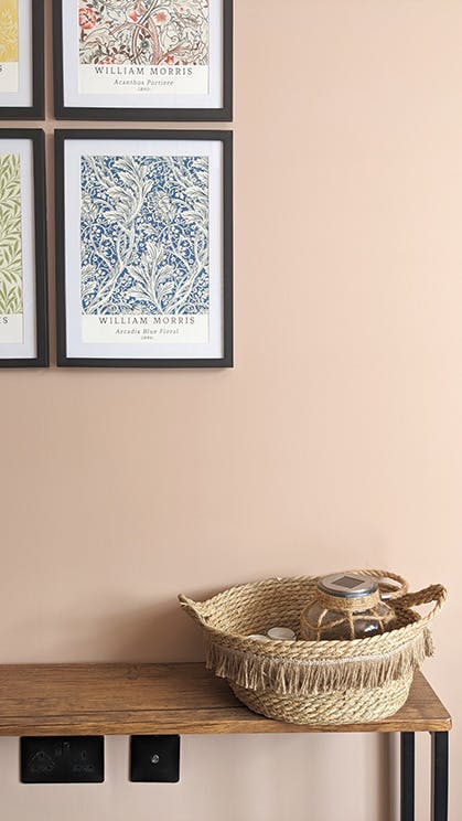 Close-up of a pink (Masquerade) wall with colorful posters in black frames and a wooden side table with a jute basket on top.
