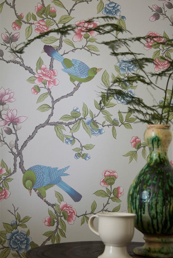 Close up of the grey floral and bird wallpaper 'Aderyn - French Grey' on a wall, with a green vase and mug on top of a table in the foreground.