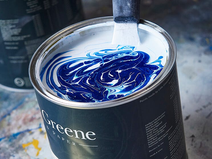 A black Little Greene paint tin containing unmixed white and blue paint, positioned on a concrete floor with paint splatters.