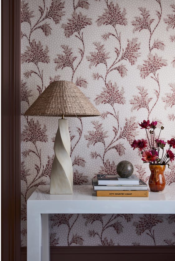 Close up of the pink floral wallpaper 'Mosaic Trail - Blush' on a wall with a white table holding a lamp, books and a vase of flowers.