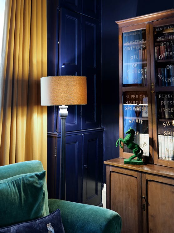 Corner of a sitting room painted with dark blue featuring a yellow curtain, a floor lamp, a green sofa and a wooden bookcase.