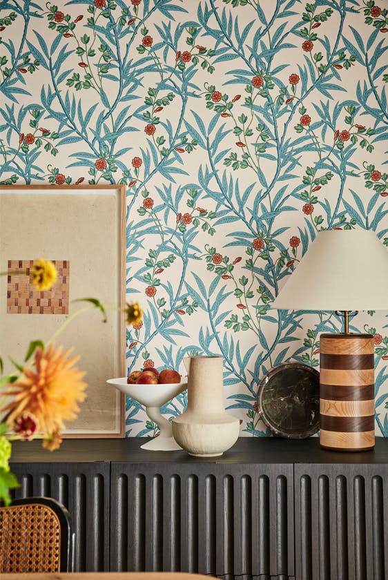 Close up of the blue and neutral floral wallpaper 'Bamboo Floral - Heat' on a wall with a dark brown radiator in the foreground with a vase, photo frame and lamp on top.