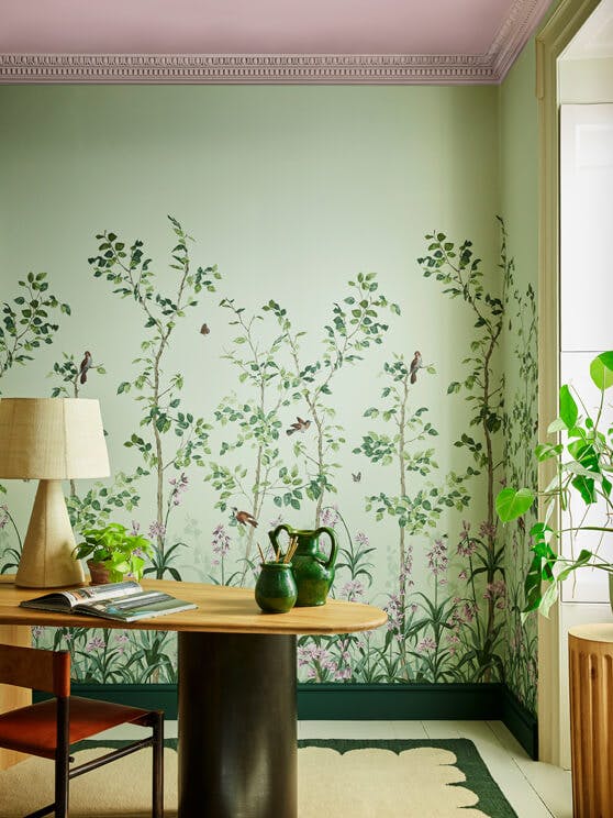 National Trust green floral mural wallpaper featuring birds and butterflies (Bird & Bluebell - Pea Green) behind a wood desk.