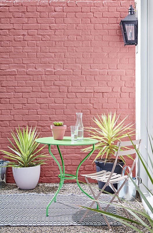 Outside brick wall painted in red shade 'Ashes of Roses' with bright green exterior table and pink chair.